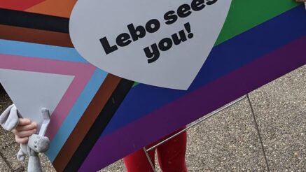 a young child holding up a yard sign that depicts the Progressive Pride Flag with a white heart in the middle that reads: Lebo sees you!